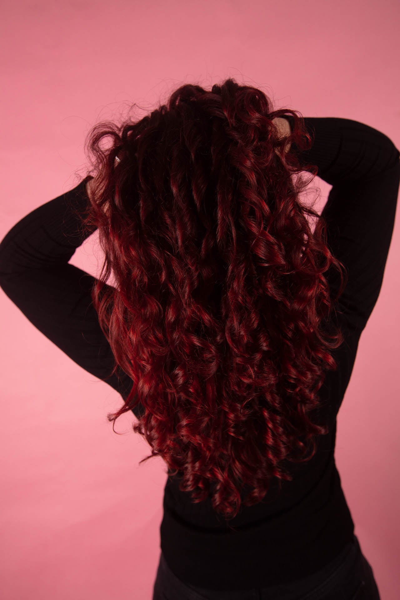 Woman running hands through beautiful deep red curly hair while standing in front of a pink backdrop